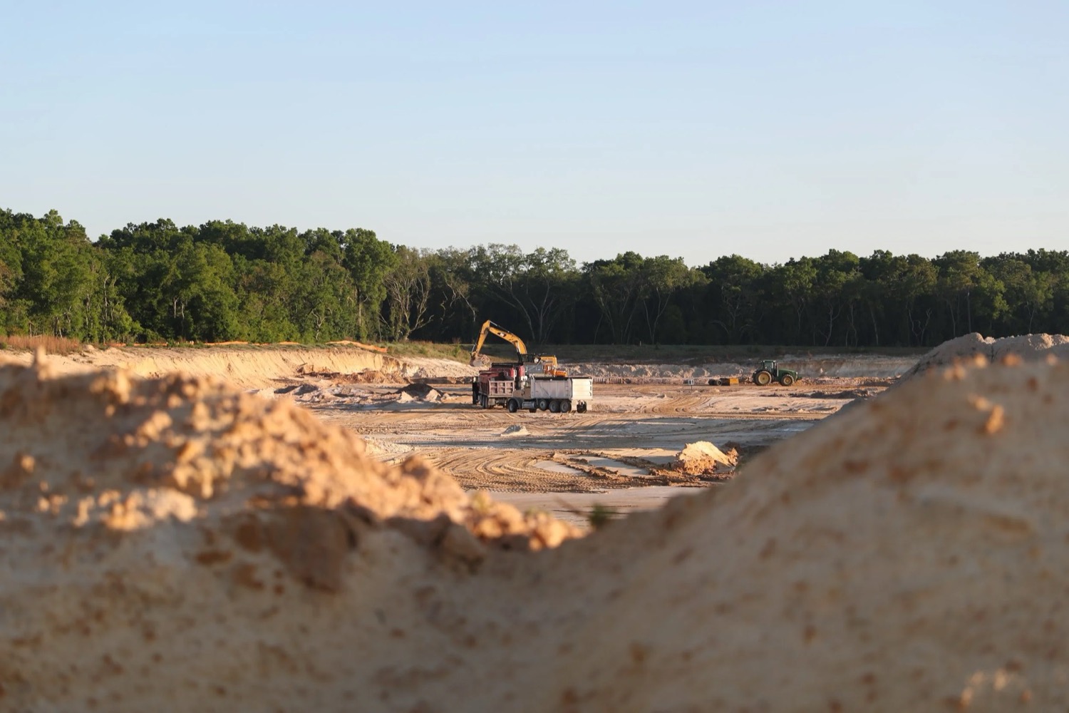 An excavator loads a dump truck in a sand pit ringed by pine trees in the late-afternoon sun